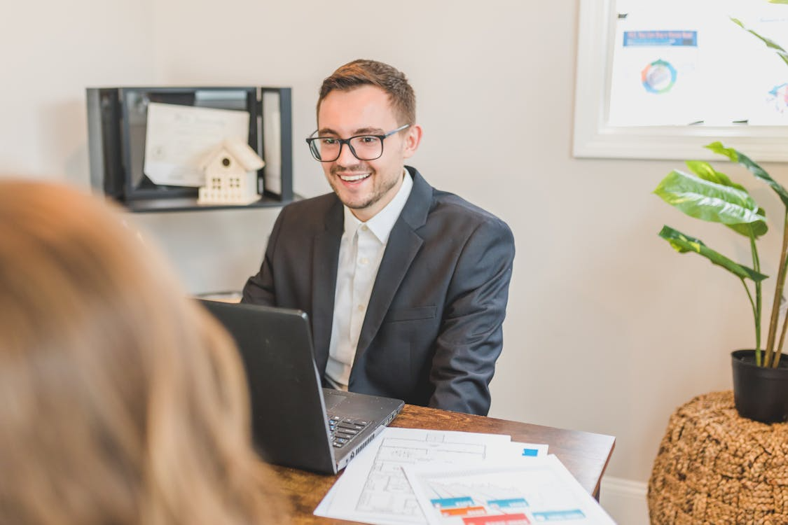 Smiling real estate agent meeting with homeowner at desk with laptop and documents.