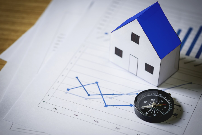 Model house on market trend charts with a compass representing analysis of factors influencing home prices.